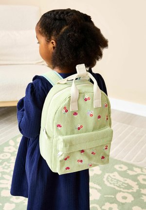 Mladá dívka s copánky, která má na sobě světle zelený manšestrový batoh zdobený malými červenými a bílými květinami, uvnitř budovy.