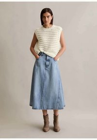 Ung kvinne står med hendene i lommene iført en ermeløs hvit strikket topp, lys blå denim midi-skjørt og brune ankelstøvler.