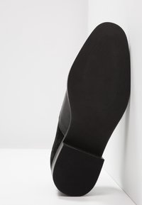 Chaussure en suède noir montrant une semelle en caoutchouc plate avec des stries texturées, inclinée contre un fond blanc pour mettre en valeur le design de la chaussure.