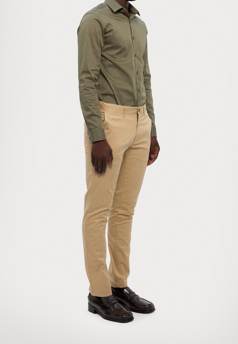 Homme portant une chemise olive à manches longues boutonnée, un pantalon beige et des chaussures de ville marron foncé, debout devant un fond uni.