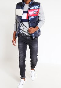 Gilet piumino navy con pannelli a blocchi di colore in crema e rosso, dotato di una patch con logo. Indossato sopra una felpa grigia chiara e jeans scuri.