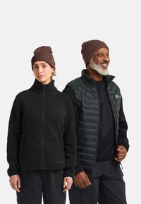 Due adulti che indossano berretti marroni e abbigliamento outdoor nero, uno con una giacca in pile e l'altro con un gilet trapuntato, in piedi uno accanto all'altro.