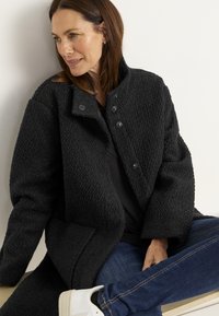 Frau mit braunen Haaren, die einen schwarzen strukturierten Mantel, dunkle Jeans und weiße Sneakers trägt, sitzt an einer weißen Wand und schaut nach links.