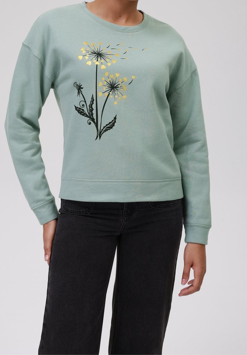 Personne portant un pull vert clair avec un motif de pissenlit noir et doré, associé à un jean noir, debout devant un fond uni.