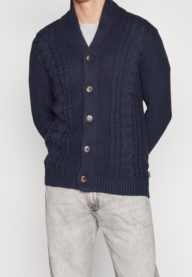 Námořnicky modrý kardigan se šálovým límcem, s pleteným vzorem copánků a pěti dřevěnými knoflíky vpředu. Nošený přes šedé tričko.