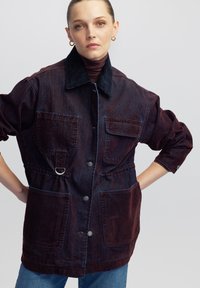 Vrouw met samengebonden haar, gekleed in gouden creolen, een donkere spijkerjack met grote zakken, een zwarte kraag en blauwe jeans, met haar handen op haar heupen.