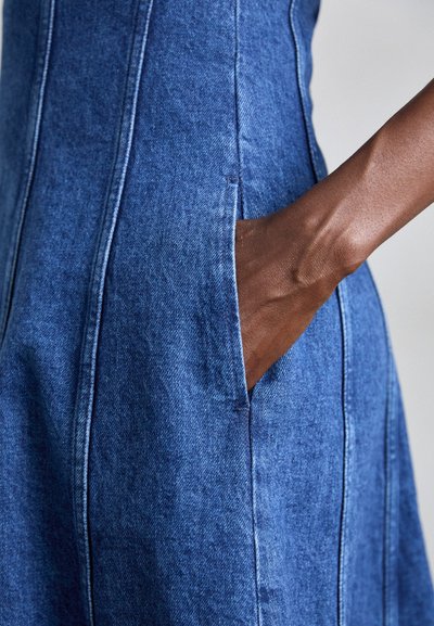 Blauwe denim jurk met verticale naden en een zijzak. De stof heeft een gestructureerde uitstraling. Hand is in de zak geplaatst.