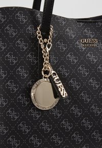 Sac à main noir de la marque Guess avec un motif répétitif du logo, chaîne en or et breloques dorées rondes et rectangulaires gravées avec les détails de la marque et de l'emplacement.