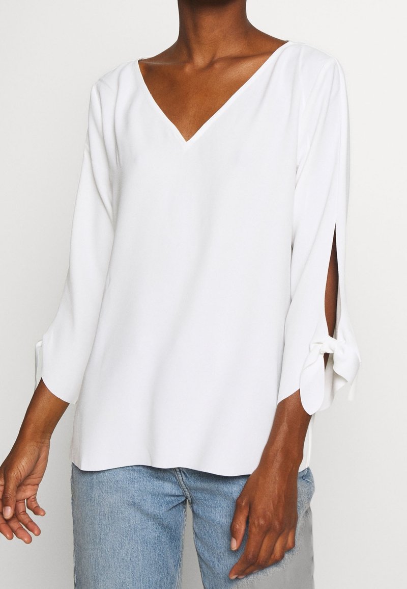 Blouse blanche avec un profond décolleté en V, manches trois-quarts avec des accents noués, et une coupe ample. Tissu doux avec une texture lisse.