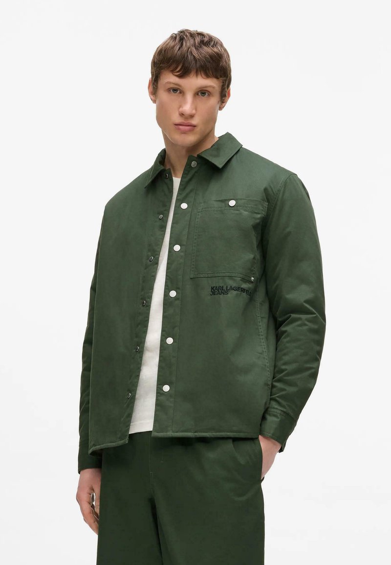 Jeune homme portant une veste-chemise boutonnée vert foncé et un pantalon assorti, avec un T-shirt blanc en dessous, debout devant un fond blanc.