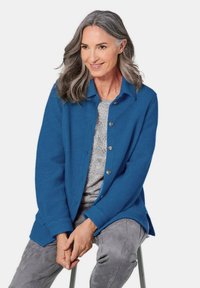 Blauwe wol cardigan met knoopsluiting, ontspannen pasvorm, lange mouwen en een spitse kraag, gecombineerd met een grijze gebreide top en grijze broek.