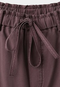 Gros plan sur la taille d’un pantalon marron avec un bandeau élastique et un cordon en tissu cousu noué en nœud à l’avant au centre.