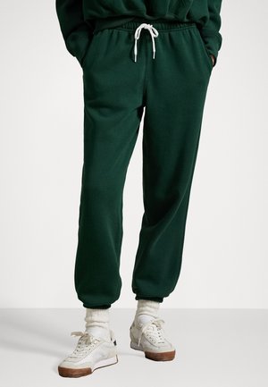 Mørkegrønne joggebukser med elastisk liv og hvit snor, med ribbet mansjetter og sidelommer, aksentuert med hvite sneakers.