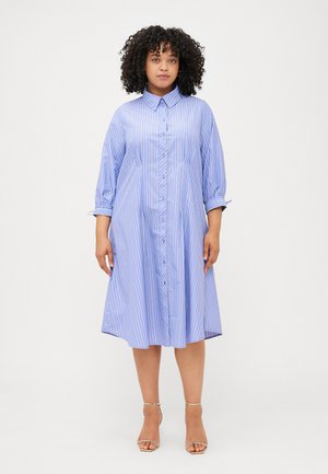 Een vrouw met krullend haar draagt een lichtblauwe gestreepte blousejurk. Ze staat zelfverzekerd en toont haar stijlvolle look met sandalen met bandjes.