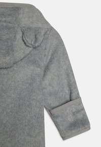 Giacca in fleece grigia con cappuccio, caratterizzata da maniche arrotolate e una texture morbida; presenta piccoli dettagli a forma di orecchie di orso sul cappuccio.