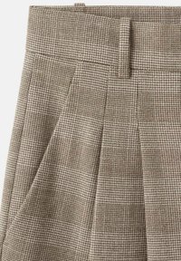 Pantalon marron à carreaux avec une ceinture ajustée, doté de plis et d'une texture en tissu finement tissé, créant un motif classique.