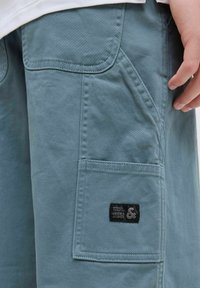 Plave pamučne cargo kratke hlače s bočnom džepom, vidljivim šavovima i crnom etiketom s "JACK & JONES" logotipom. Gladak materijal, ležeran dizajn.