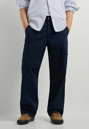 Persona con pantalones cargo azul oscuro, zapatillas marrones y una camisa de rayas con botones remangada, con las manos en los bolsillos.