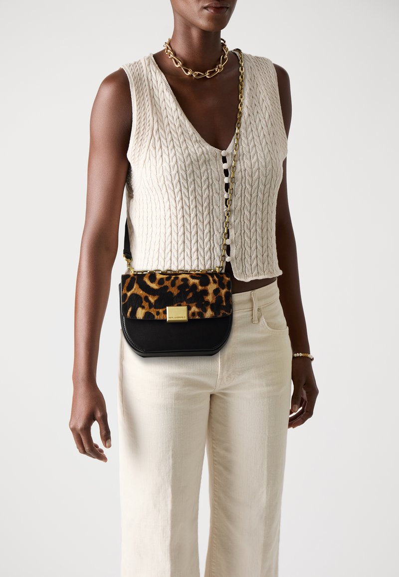 Leopard print crossbody bag with a black base, gold hardware, and a chain strap, paired with a cream knitted vest and light trousers.
