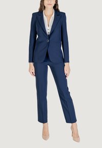 Completo blu navy su misura composto da un blazer aderente e pantaloni slim, abbinato a una maglietta bianca. Tessuto morbido con un leggero lucido.
