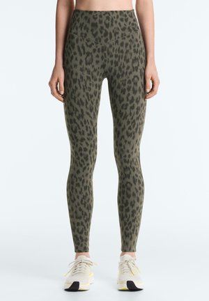 Kvinde iført højtaljede olivengrønne leggings med sorte leopardpletter og hvide sneakers, stående mod en ensfarvet baggrund.