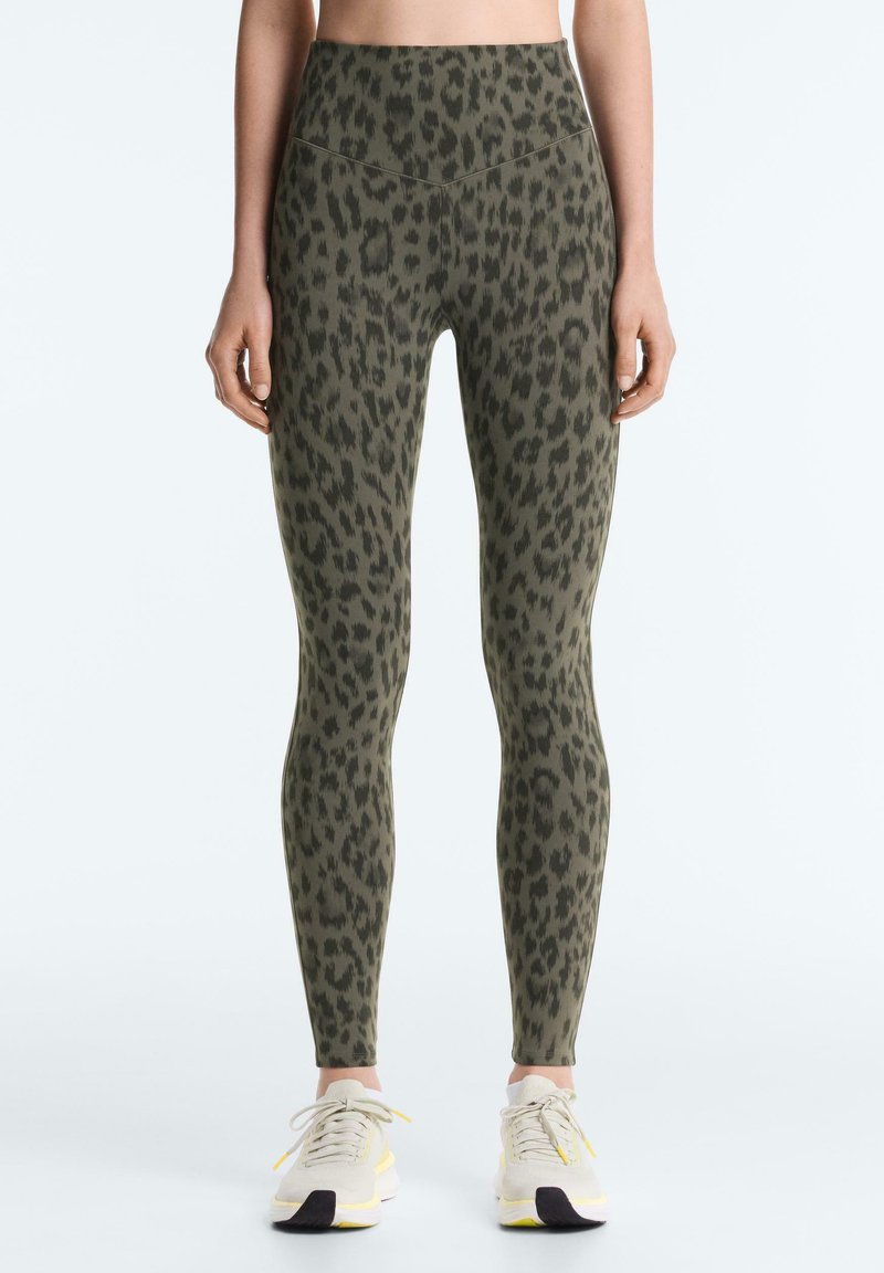 Femme portant des leggings taille haute vert olive avec des taches léopard noires et des baskets blanches, debout devant un arrière-plan uni.