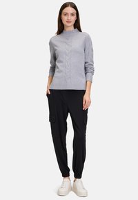 Grauer gestrickter Pullover mit einem gerippten Kragen und Zopfmuster, kombiniert mit schwarzen Cargo-Hosen und weißen Sneakers, alles vor einem einfarbigen Hintergrund.