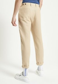 Pantaloni di lino beige con risvolti, due tasche posteriori e una texture liscia, abbinati a sneakers bianche e calzini con dettagli blu.
