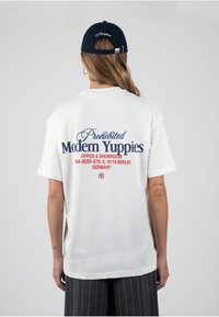 Άτομο που φοράει λευκό T-shirt με το κείμενο «Prohibited Modern Yuppies» και διεύθυνση, σε συνδυασμό με σκούρο παντελόνι με ρίγες και σκούφο ναυτικού μπλε που φοράει ανάποδα.