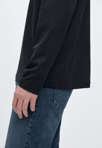 Svart långärmad skjorta med en jämn textur, kombinerad med blå jeans. Skjortan har en rundad nederkant och åtsittande ärmar.