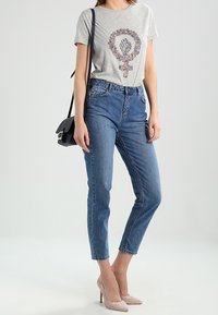 Maglietta grigia con simbolo femminista floreale, jeans blu a vita alta e décolleté rosa chiaro. Piccola borsa a tracolla nera con cinturino.