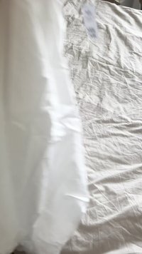 Biała narzuta wykonana z pomarszczonego materiału, częściowo nałożona na pomarszczony beżowy prześcieradło. Na brzegu widoczna etykieta z instrukcją pielęgnacji.
