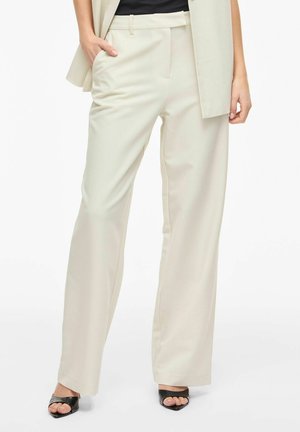 Femme portant un pantalon beige à jambes larges, un haut noir, un blazer beige et des talons noirs à bout ouvert, debout avec une main dans la poche.