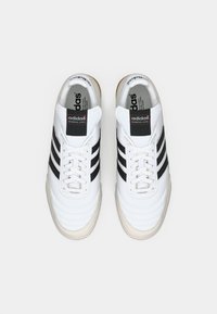 Para białych butów piłkarskich Adidas Mundial Goal z czarnymi paskami,wiązania związane, widok z góry na jednolitym tle.