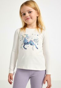 Langærmet hvid top med et fantasifuldt unicorn-design i multikolorede palietter, sammen med teksturerede lyserøde leggings.