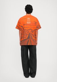 Orange kortærmet T-shirt med et vej- og bjergmotiv. Har hvid tekst på ryggen. Sættes sammen med sorte brede cargo-bukser.