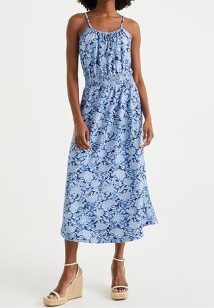 Kobieta w niebieskiej sukience midi w kwiaty z cienkimi ramiączkami i marszczonym stanem, w połączeniu z beżowymi sandałami na koturnie.