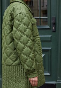 Chaqueta acolchada color verde oliva con un patrón de diamante, textura acanalada contrastante en la parte inferior y detalle de puños con botones a presión.