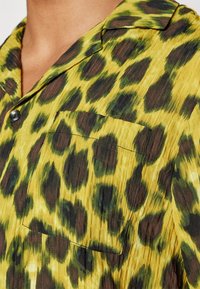 Camicia gialla con macchie leopardate nere e verdi, tessuto testurizzato, tasca sul petto anteriore e bottone nero vicino al colletto.