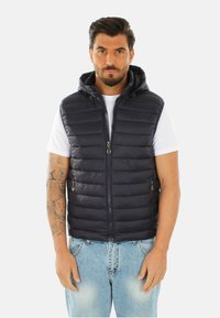 Gilet imbottito blu navy con cappuccio; design trapuntato; cerniera frontale con due tasche con zip; indossato sopra una maglietta bianca e jeans chiari.
