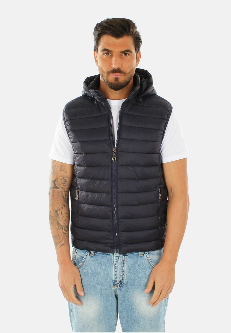 Gilet imbottito blu navy con cappuccio; design trapuntato; cerniera frontale con due tasche con zip; indossato sopra una maglietta bianca e jeans chiari.