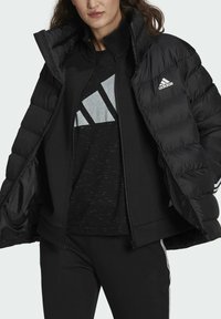 Doudoune noire à col montant, équipée d'une fermeture éclair et du logo Adidas. Portée sur un sweat-shirt noir avec un logo argenté.