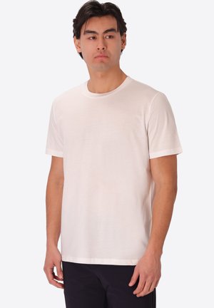 Uomo che indossa una semplice t-shirt bianca a maniche corte con scollo rotondo e pantaloni scuri, che guarda di lato su uno sfondo chiaro uniforme.