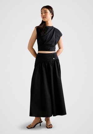 Vrouw staand met handen achter de rug, gekleed in een zwarte mouwloze cropped top, een lange high-waisted rok met gesp, en zwarte sandalen met hak.