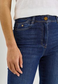 Los vaqueros de mezclilla azul oscuro presentan un diseño de talle alto, detalles en hardware dorado y una textura suave con un ligero desgaste y detalles de difuminado.