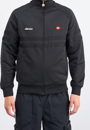 Osoba ubrana w czarną kurtkę ellesse na zamek z naszywkami z logo na piersi i ciemne spodnie z kieszeniami i zamkami.