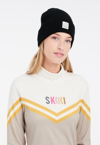 Schwarze gerippte Mütze mit einem Logo-Patch, getragen zu einem hellen Oberteil mit gelb-rosafarbenen Streifenmustern. Glatte Stofftextur.