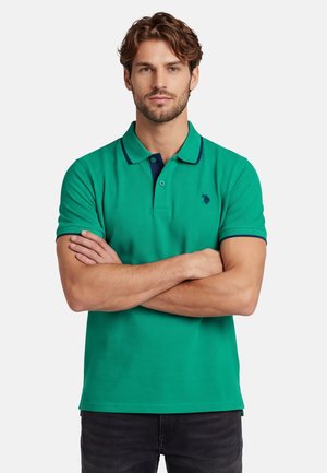 Mann mit verschränkten Armen, der ein grünes Polo mit marineblauem Besatz und kleinem marineblauem Logo auf der linken Brust vor einem einfarbigen Hintergrund trägt.