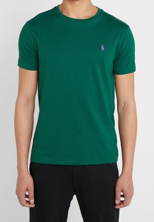 Homme portant un t-shirt vert ajusté à manches courtes avec un petit logo brodé violet sur la poitrine gauche, associé à un pantalon noir.