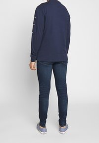 Mörkblå långärmad tröja med text på ärmarna, kombinerad med blå jeans och grå sneakers. Slät tyg, avslappnad design, figurvänlig stil.